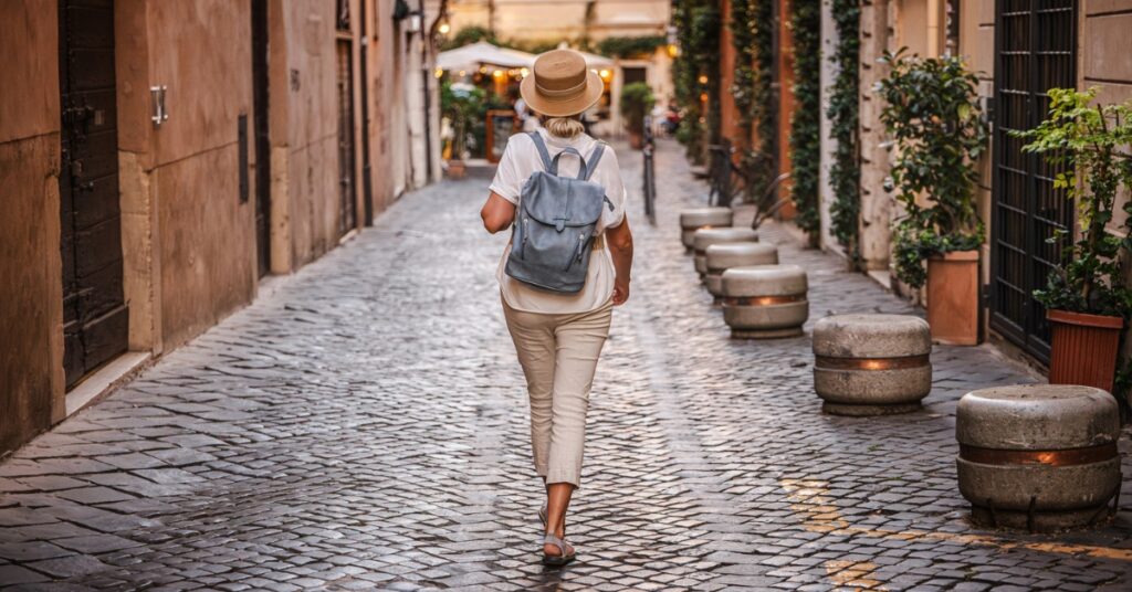 Woman in a sun hat and backpack walking along a cobbled street lined with plants and old buildings
