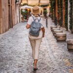 Woman in a sun hat and backpack walking along a cobbled street lined with plants and old buildings